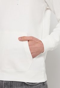 Weißer Sweatshirt mit Kängurutasche, gerippten Bündchen und Taillenbund. Weicher Stoff und glatte Oberfläche, zeigt eine Hand, die in der Tasche ruht.