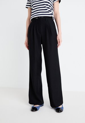 Schwarze, weit geschnittene Hose mit hoher Taille und einer Vorderfalten-Design. Kombiniert mit einem navy-weißen gestreiften T-Shirt und blauen Ballerinas.