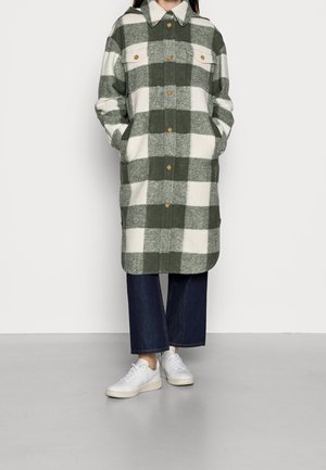Personne portant un long manteau boutonné à carreaux verts et blancs, un pantalon large bleu foncé et des baskets blanches, debout les mains dans les poches.