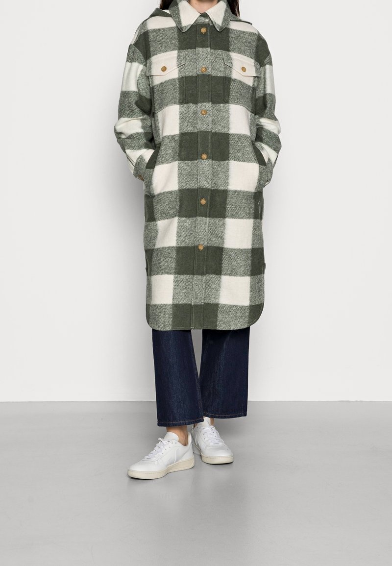 Personne portant un long manteau boutonné à carreaux verts et blancs, un pantalon large bleu foncé et des baskets blanches, debout les mains dans les poches.