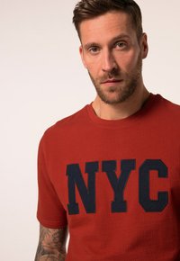 Červené tričko s texturou, které na přední straně zdobí výrazné námořnické modré písmo "NYC". Kulatý výstřih a krátké rukávy.