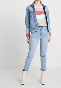 Donna che indossa un giubbotto in denim azzurro chiaro e jeans, un maglione a righe pastello e stivaletti beige che si trova davanti a uno sfondo neutro.