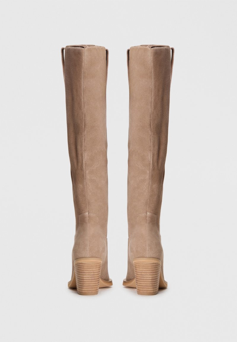 Un par de botas altas de ante beige con tacones de madera apilados, mostradas desde atrás sobre un fondo blanco liso.