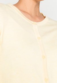 Jasnożółty kardigan z okrągłym dekoltem, zapinany na guziki z przodu, wykonany z fakturowanej dzianiny. Sześć pasujących guzików wzdłuż pliski.