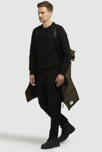 Maglione nero, pantaloni cargo neri e stivali neri, con un giubbotto verde oliva indossato aperto e drappeggiato sulla spalla.