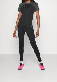 Czarny koszulka sportowa z teksturowanymi szarymi ramionami, w połączeniu z czarnymi legginsami z geometrycznym wzorem i pomarańczowym logo Nike; różowe buty sportowe.