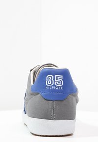 Baskets grises avec un accent en cuir bleu sur le talon. Présentent le "85" en blanc à l'arrière, une texture en toile et une semelle en caoutchouc blanc.