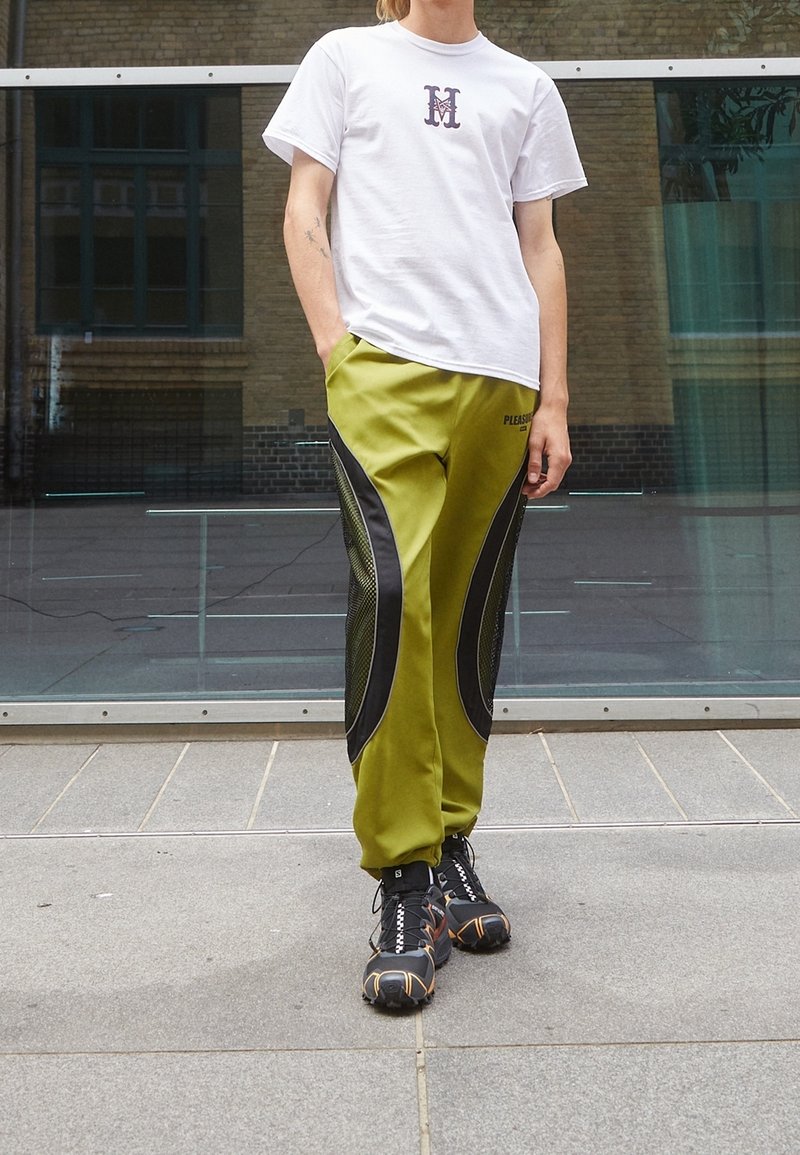 T-shirt blanc avec logo violet, associé à un pantalon vert olive avec des accents en maille noire. Baskets noires avec des détails orange.