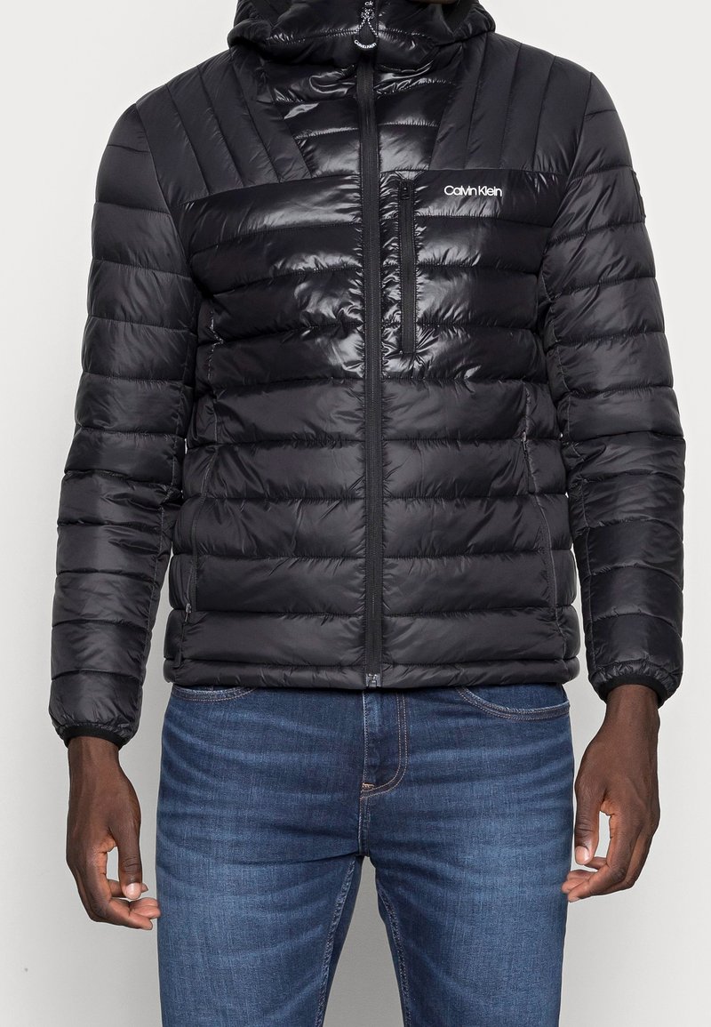 Zwarte donsjas met capuchon, voorzien van een horizontaal doorgestikt ontwerp, ritssluiting aan de voorkant en een borstzak met merklogo. Getextureerde glanzende afwerking.