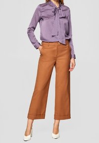 Ljuslila blus med mönster och rosett i halsen, kombinerad med kamelfärgade byxor med vida ben, avslutad med vita klackskor.