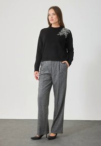 Zwarte trui met een gestructureerd patroon en zilveren versiering op de schouder, gecombineerd met grijze geplooide broek en zwarte hoge hakken.