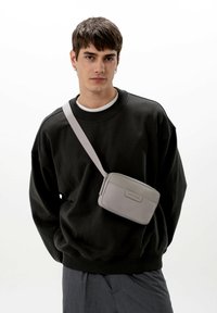 Šedá crossbody taška vyrobená z hladké kůže, obdélníkového tvaru, s přední nášivkou a plochým popruhem nošeným přes černou mikinu.