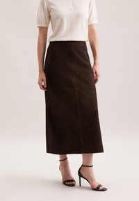 Jupe midi en daim marron, avec une coupe droite et des coutures visibles. Associée à un haut de couleur claire et des sandales à talons noires.