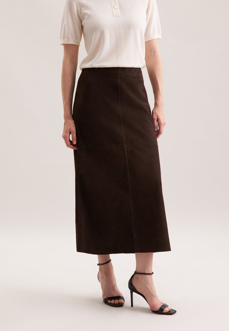 Jupe midi en daim marron, avec une coupe droite et des coutures visibles. Associée à un haut de couleur claire et des sandales à talons noires.