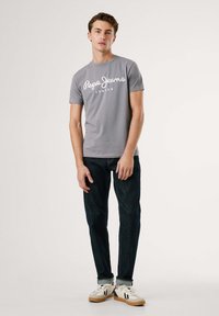 Šedé tričko s krátkým rukávem a bílým logem "Pepe Jeans London", spárováno s tmavými džíny a bílými teniskami. Jednoduché pozadí s jemným osvětlením.