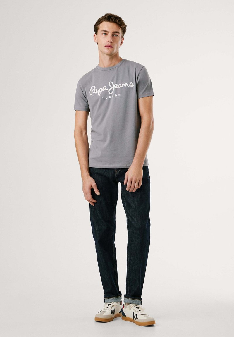 Graues T-Shirt mit kurzen Ärmeln und weißem "Pepe Jeans London" Logo, kombiniert mit dunklen Jeans und weißen Turnschuhen. Schlicher Hintergrund mit sanfter Beleuchtung.