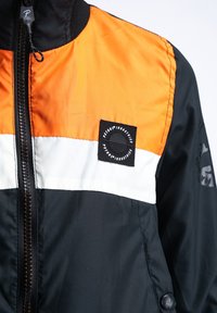 Zwarte, oranje en witte windbreaker jas met een ritsluiting, geribde kraag en een embleem met een logo op de borst, gladde textuur.