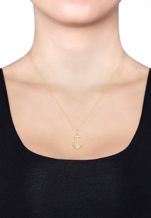 Goldener Anker-Anhänger an einer feinen Kette, vor einem schwarzen Oberteil. Das Anker-Design weist spitze Kanten und eine glatte Oberfläche auf.