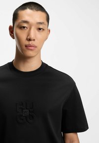 Camiseta de algodón negra con un logo en relieve en el centro, cuello redondo y mangas cortas con una textura suave.