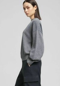 Felpa grigia in maglia con spalle scese, polsini e orlo a costine, indossata con pantaloni cargo scuri. Tessuto liscio, vestibilità rilassata, marchio minimale.