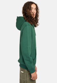 Sudadera con capucha verde y bolsillo frontal, hecha de una tela suave. Presenta un corte relajado y puños y dobladillo acanalados.
