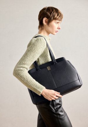 Kobieta z krótkimi włosami, ubrana w jasnozielony sweter o fakturze i czarne spodnie, niosąca na ramieniu dużą czarną torbę ze skóry.