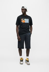 Μαύρο βαμβακερό T-shirt με πολύχρωμο γραφικό "Air Jordan", συνδυασμένο με σκούρο σορτς και αθλητικά παπούτσια με χρώματα μπεζ, κίτρινο και μαύρο.