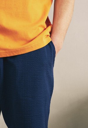 Person iført strukturerede marineblå bukser og en skrigorange skjorte, med den ene hånd afslappet i lommen mod en ensfarvet baggrund.