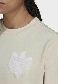 Light beige sweatshirt featuring a textured white fringe logo with three leaf shapes and horizontal stripes. Soft fabric and relaxed fit.