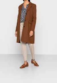 Brauner Wollmantel mit Revers und drei Knöpfen, kombiniert mit einer blauen gemusterten Bluse und beigen, taillierten Hosen. Braune Slipper runden das Outfit ab.