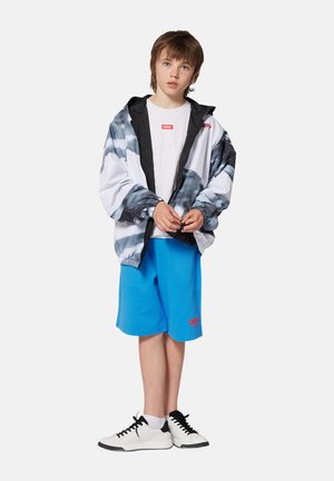 Veste coupe-vent noire et blanche, shorts bleus et baskets blanches avec des accents noirs. La veste dispose d'une fermeture éclair et de poignets élastiques.