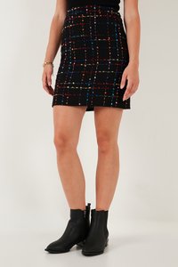 Donna che indossa una minigonna nera in tweed con motivo a griglia multicolore e stivaletti neri, in piedi davanti a uno sfondo semplice.