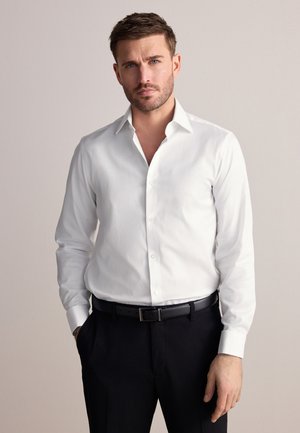 Man met kort bruin haar en baard, gekleed in een wit overhemd en zwarte broek, staand met één hand in de zak tegen een effen achtergrond.