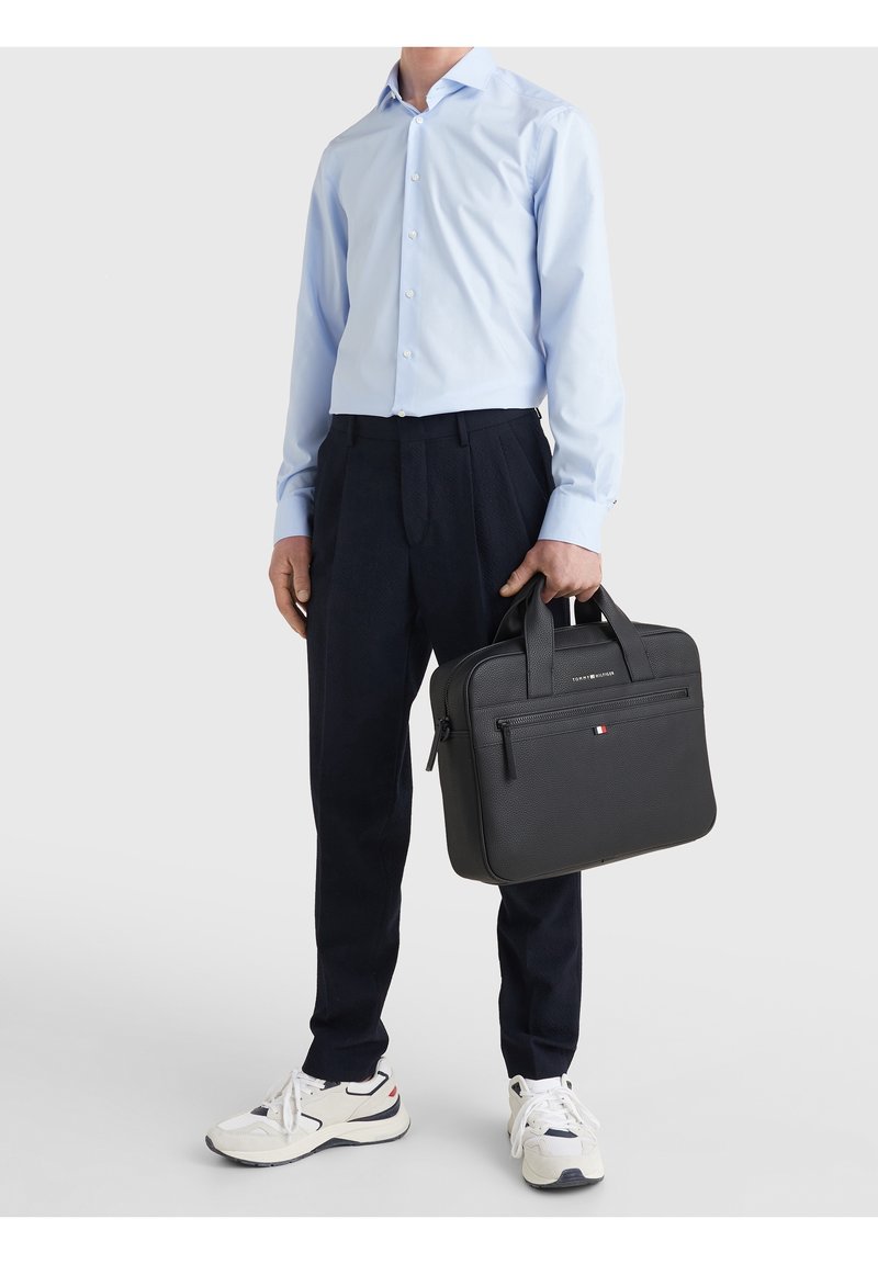 Homme portant une chemise bleu clair, un pantalon noir et des baskets blanches, tenant une mallette en cuir noire avec une poche zippée à l'avant.