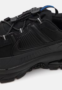 Svart sneaker med en teksturert overdel med horisontale striper, gummisåle og elastiske snører med en glideknapp for en sikker passform.