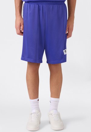 Männliche Person trägt blaue Mesh-Sportshorts, weiße Crew-Socken und weiße Turnschuhe und steht vor einem schlichten hellen Hintergrund.