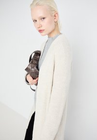 Krema pletena kardigan z odprtim sprednjim delom, nošen preko sivega puloverja s turtleneck izrezom. Model drži temno rjav čevljek, kar prikazuje teksture in nevtralne barve.