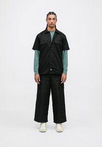 Muž s opletenými vlasy stojí v černém tričku s krátkým rukávem přes zelené dlouhé rukávy, v černých širokých prošívaných kalhotách a v bílých teniskách.