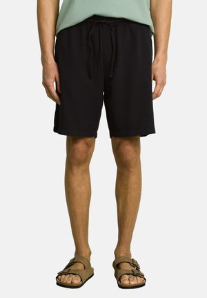 Person trägt schwarze Shorts mit Kordelzug und braune Sandalen mit Schnalle und steht vor einem schlichten Hintergrund.
