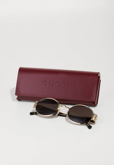 Gafas de sol Gucci con lentes marrones, montura de metal dorado y puntas de las patillas negras. Acompañadas de un estuche de cuero burdeos con el logo "GUCCI" grabado.