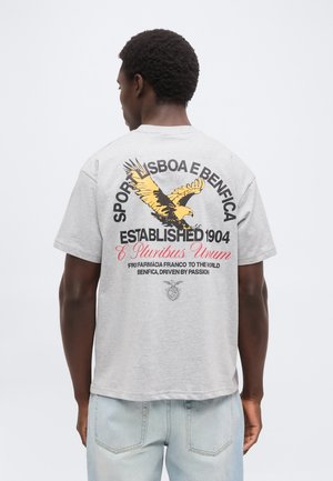 Mann trägt ein graues T-Shirt mit einem gelben Adler und dem Text „Sport Lisboa e Benfica Established 1904 E Pluribus Unum“ auf dem Rücken.