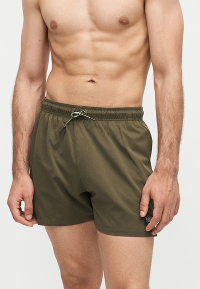 Μυώδης ανδρικός κορμός που φοράει ελαιώδη πράσινα μαγιό με ελαστική μέση και κορδόνι, στέκεται μπροστά σε ένα απλό λευκό φόντο.