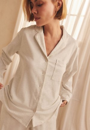 Femme aux cheveux courts portant un haut beige clair à manches longues boutonné avec col et poche poitrine, debout devant des rideaux translucides.