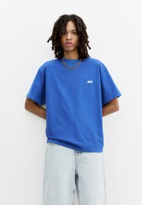 Blå bomulds t-shirt med en afslappet pasform, der har korte ærmer og et lille hvidt logo på brystet, parret med lyse denimshorts.