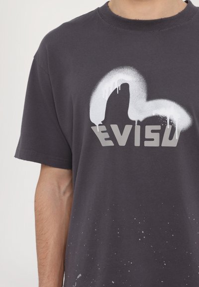 Camiseta de algodón gris oscuro con un diseño pintado en blanco y la palabra "EVISU" en una fuente gruesa y en bloque, con acentos de salpicaduras.