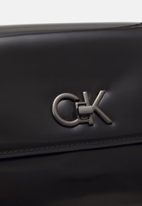Primo piano di una borsa nera con finitura lucida e un logo CK in evidenza in hardware tono argento. Design sofisticato e moderno.