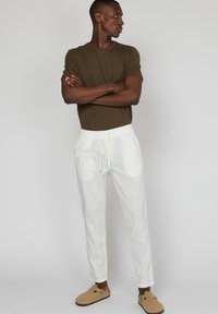 Camiseta de manga corta verde oliva, pantalones blancos con cordón y zapatos marrones tipo slip-on. El atuendo presenta un corte relajado y una textura suave.