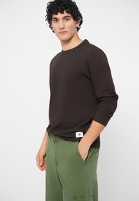 Smeđi dukserica dugih rukava s okruglim izrezom, teksturirani materijal i mala bijela oznaka, u kombinaciji s zelenim cargo hlačama s bočnim džepovima.