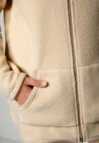 Chaqueta de forro polar beige con cierre frontal, dos bolsillos laterales y tejido texturizado. El puño tiene un acabado suave y redondeado.