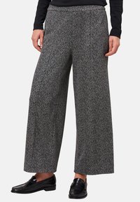 Persona che indossa pantaloni larghi grigi a motivo houndstooth con mocassini in pelle nera e una maglia nera a maniche lunghe, in piedi davanti a uno sfondo neutro.